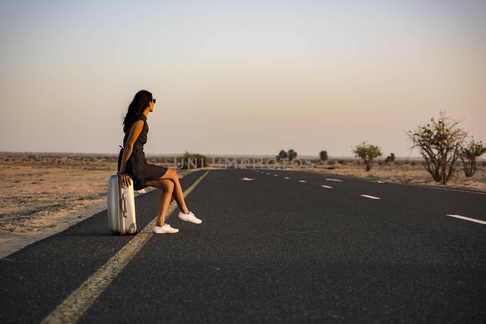 Woman waiting with her luggage on rural road in the desert for a lift or a vehicle to come