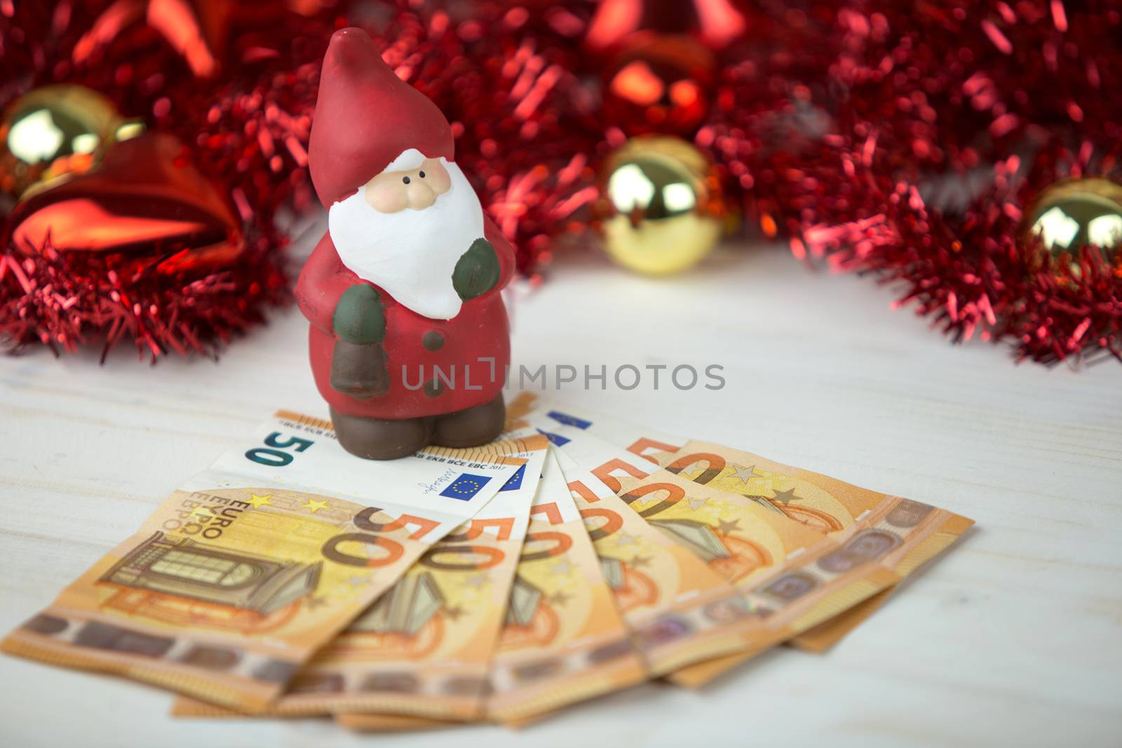 Christmas money business concept: a statuette of Santa Claus on some fifty euro banknotes with red and gold baubles and wreath decoration with bokeh effect on light wooden table