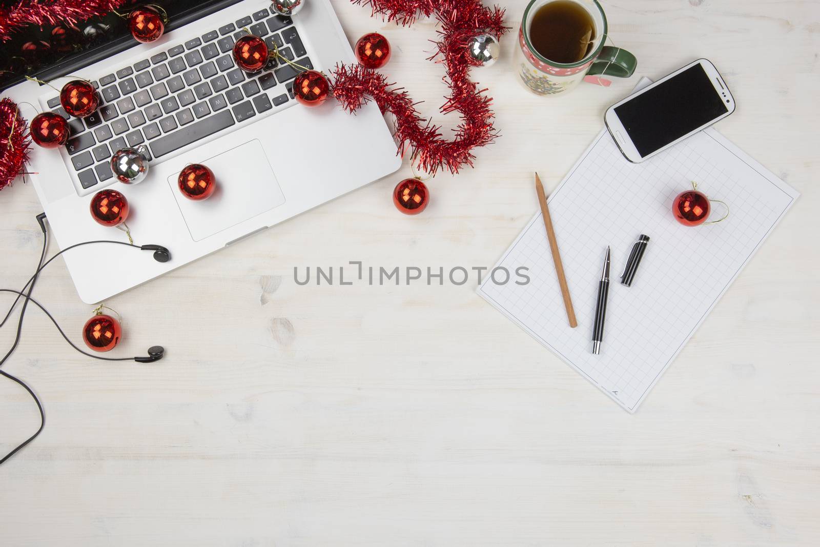 Job at Christmas holidays: flat lay of a light wooden table with an open aluminum laptop, red decoration, red baubles, Christmas cup of tea, smartphone, block notes, pencil and pen on block note by robbyfontanesi