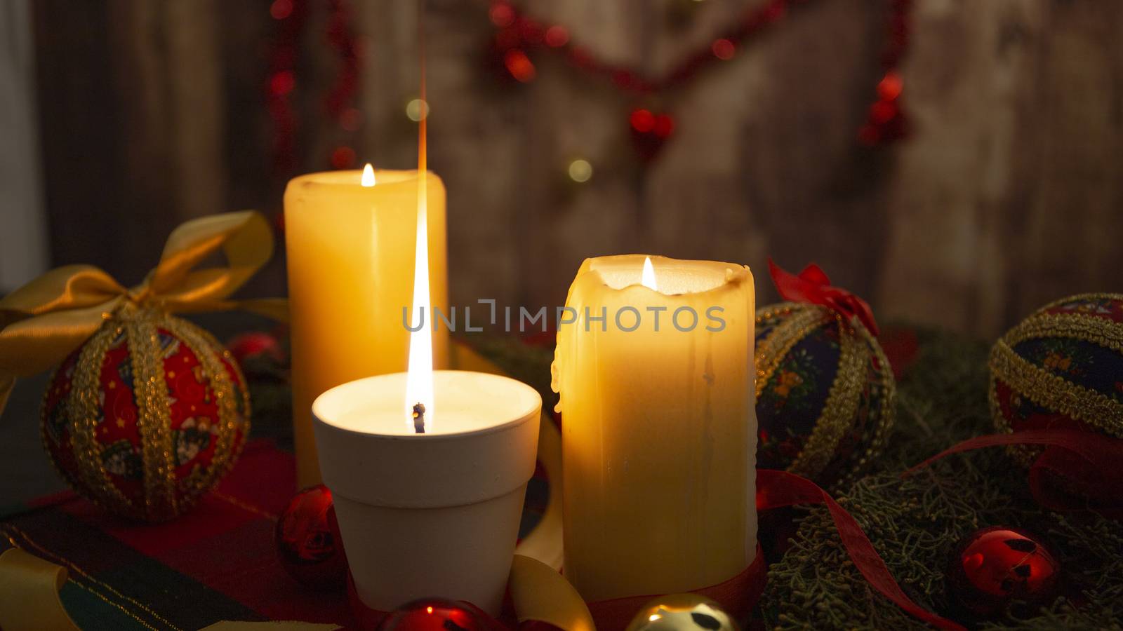 Close up of a Lit candle with big flame on Christmas table cloth with around pine branches, decoupage baubles, with lit candles and hanging Christmas decoration on wooden background with bokeh effect