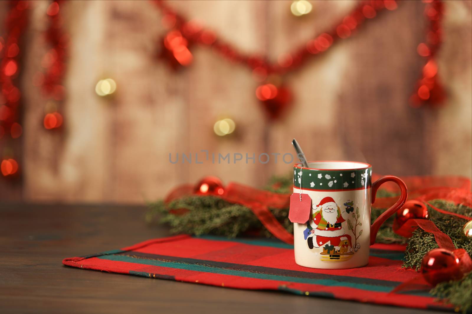 Christmas mug with hanging empty tea label on Christmas table cloth with around pine branches, red baubles and hanging Christmas decoration on wooden background with bokeh effect by robbyfontanesi