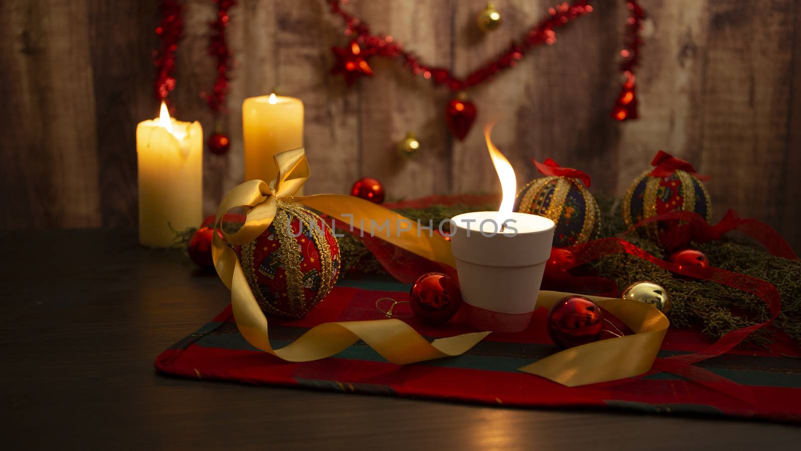 Lit candle with big flame on Christmas table cloth with around pine branches, decoupage baubles, with lit candles and hanging Christmas decoration on wooden background with bokeh effect