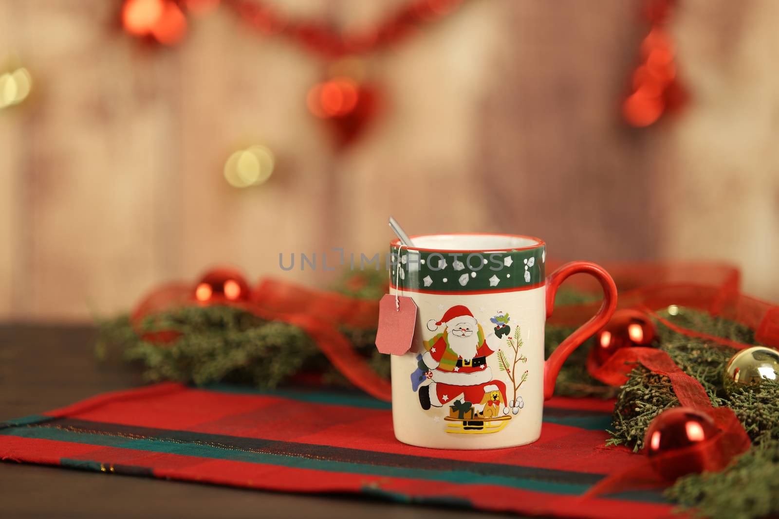 Close up of a Christmas mug with hanging empty tea label on Christmas table cloth with around pine branches, red baubles and hanging Christmas decoration on wooden background with bokeh effect by robbyfontanesi
