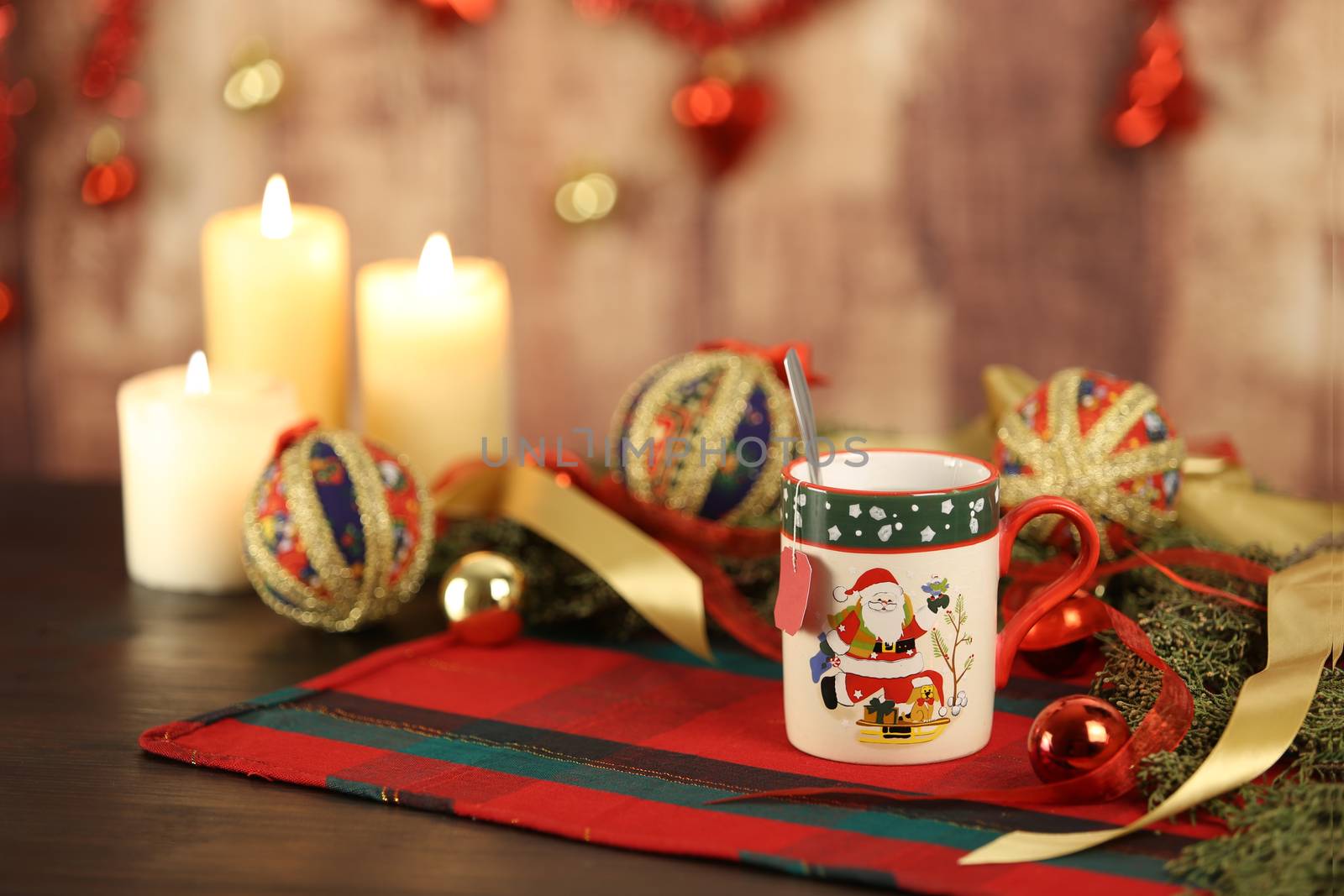 Christmas mug with empty tea label on Christmas table cloth with around pine branches, decoupage baubles, with lit candles and hanging Christmas decoration on wooden background with bokeh effect