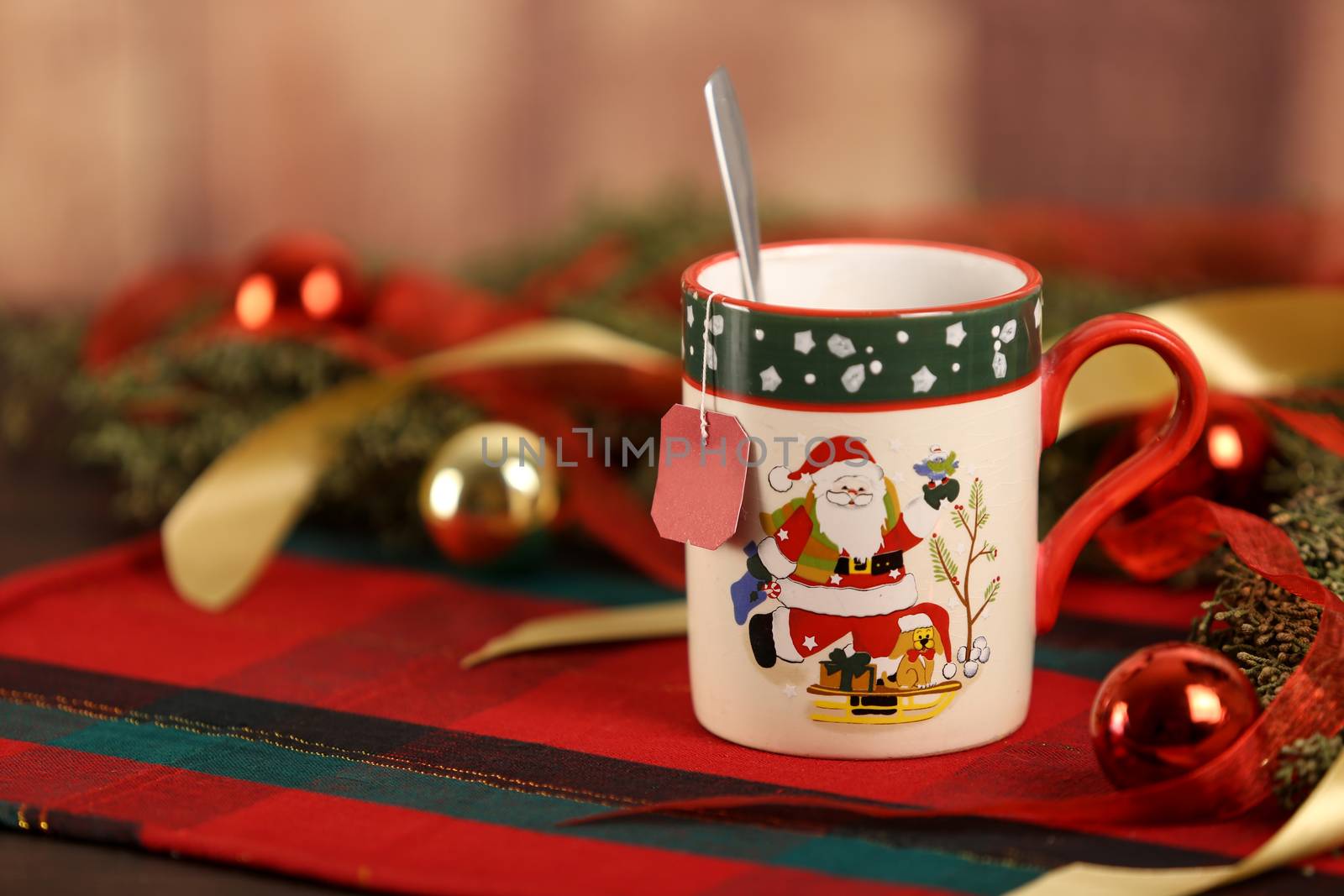 Close up of a Christmas mug with hanging empty tea label on Christmas table cloth with around pine branches, red baubles and hanging Christmas decoration on wooden background with bokeh effect