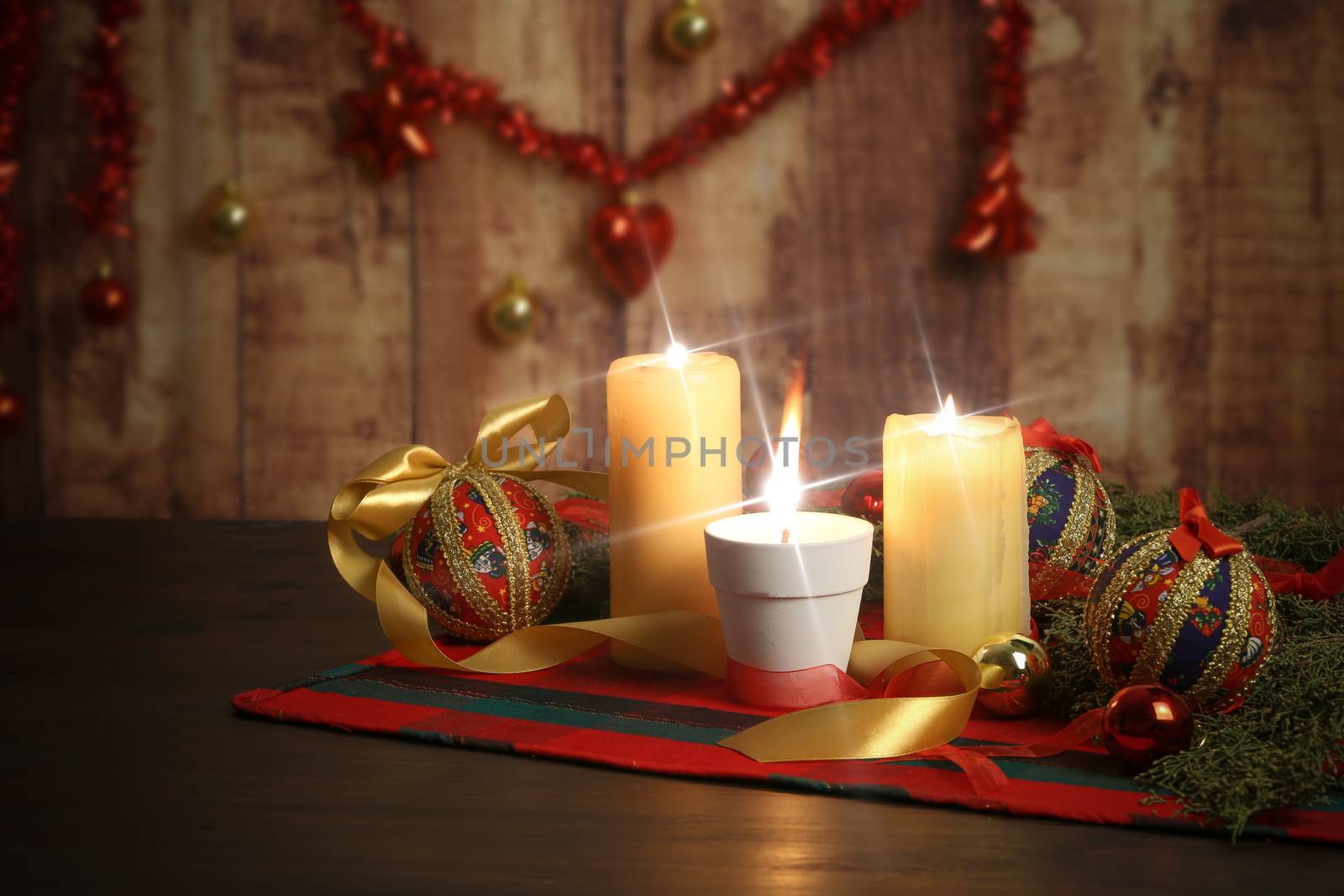 Lit candle with big flame on Christmas table cloth with around pine branches, decoupage baubles, with lit candles and hanging Christmas decoration on wooden background with bokeh effect