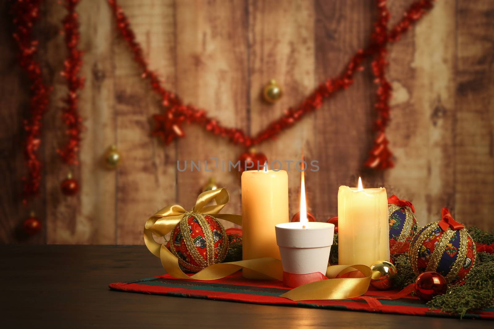 Lit candle with big flame on Christmas table cloth with around pine branches, decoupage baubles, with lit candles and hanging Christmas decoration on wooden background with bokeh effect