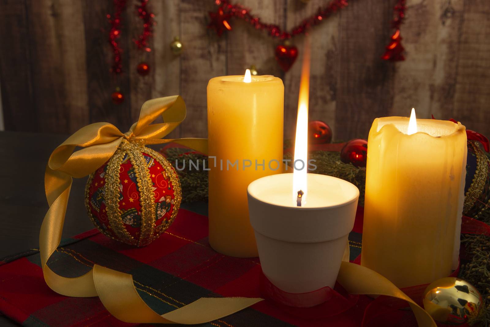 Close up of a Lit candle with big flame on Christmas table cloth with around pine branches, decoupage baubles, with lit candles and hanging Christmas decoration on wooden background with bokeh effect by robbyfontanesi