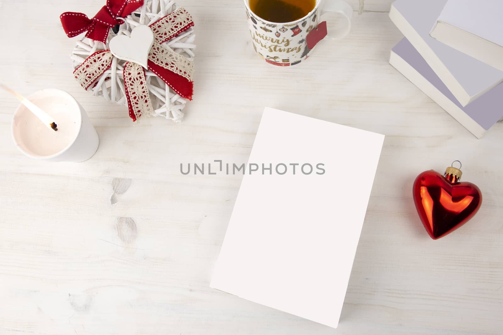 Christmas view from top for a book presentation: book with blank cover with a white lit candles, Christmas decorated cup of tea, red heart bauble, a shabby decorated white heart on light wooden table by robbyfontanesi