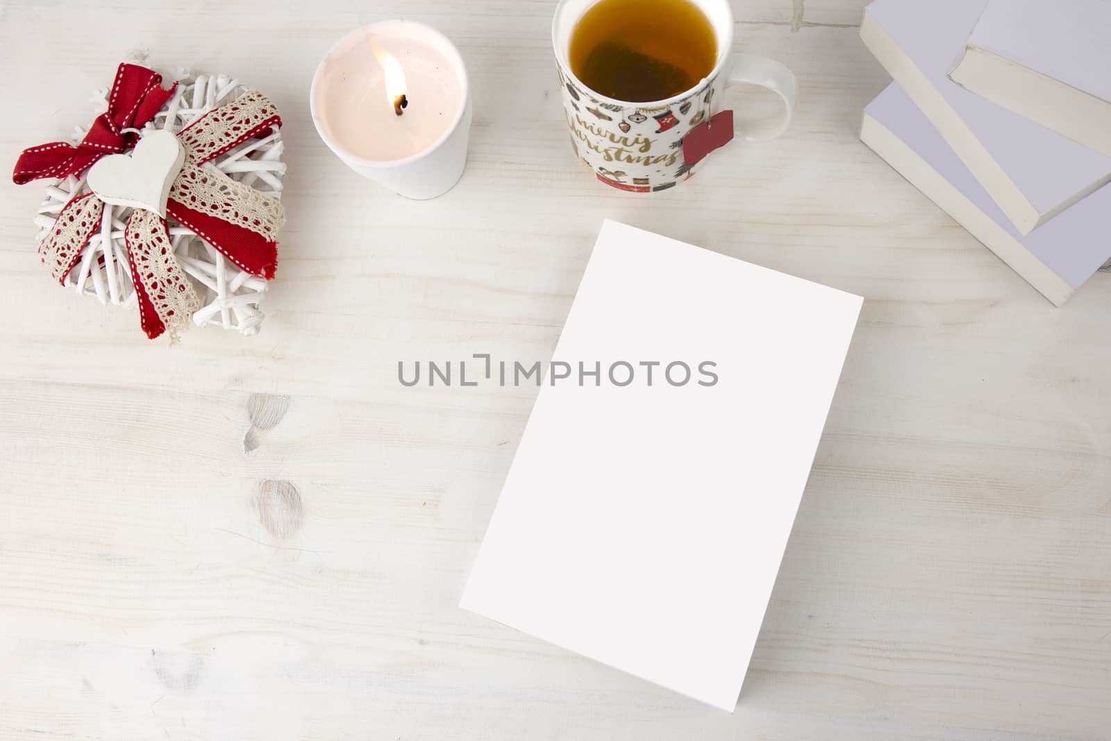 Christmas view from top for a book presentation: book with blank cover with a white lit candles, Christmas decorated cup of tea other three books, a shabby decorated white heart on light wooden table by robbyfontanesi