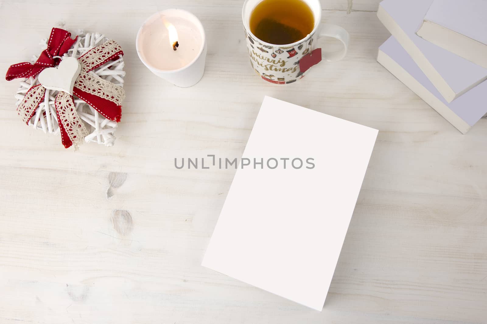 Christmas view from top for a book presentation: book with blank cover with a white lit candles, Christmas decorated cup of tea other three books, a shabby decorated white heart on light wooden table