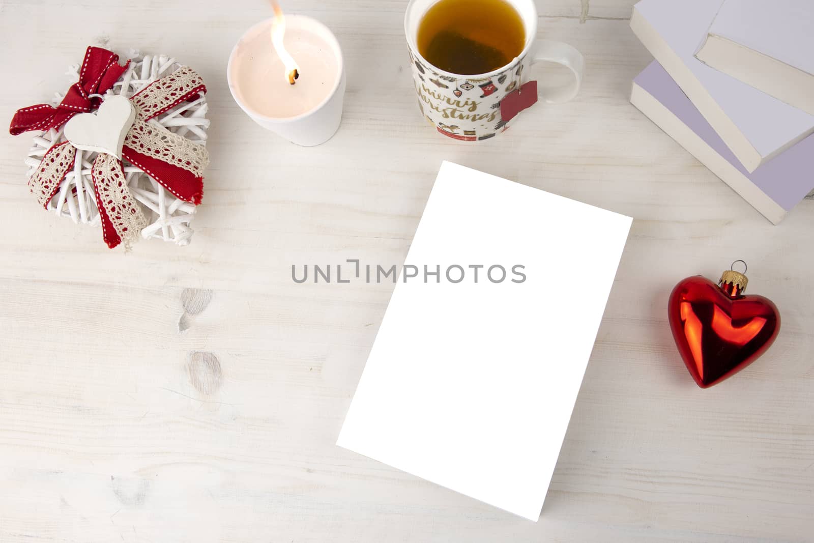 Christmas view from top for a book presentation: book with blank cover with a white lit candles, Christmas decorated cup of tea, red heart bauble, a shabby decorated white heart on light wooden table