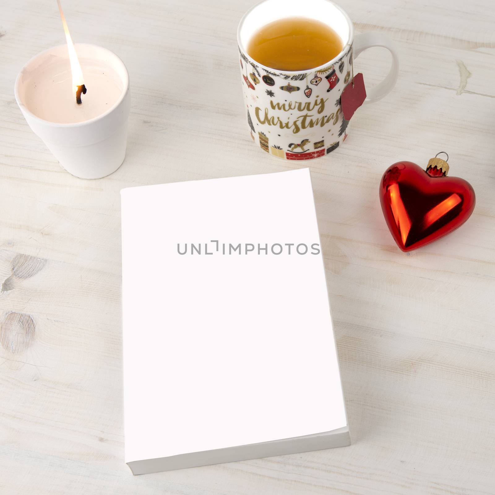 Christmas setting view from top for a book presentation: book with blank cover with a white lit candles, Christmas cup of tea and red baubles with heart shape on light wooden table by robbyfontanesi