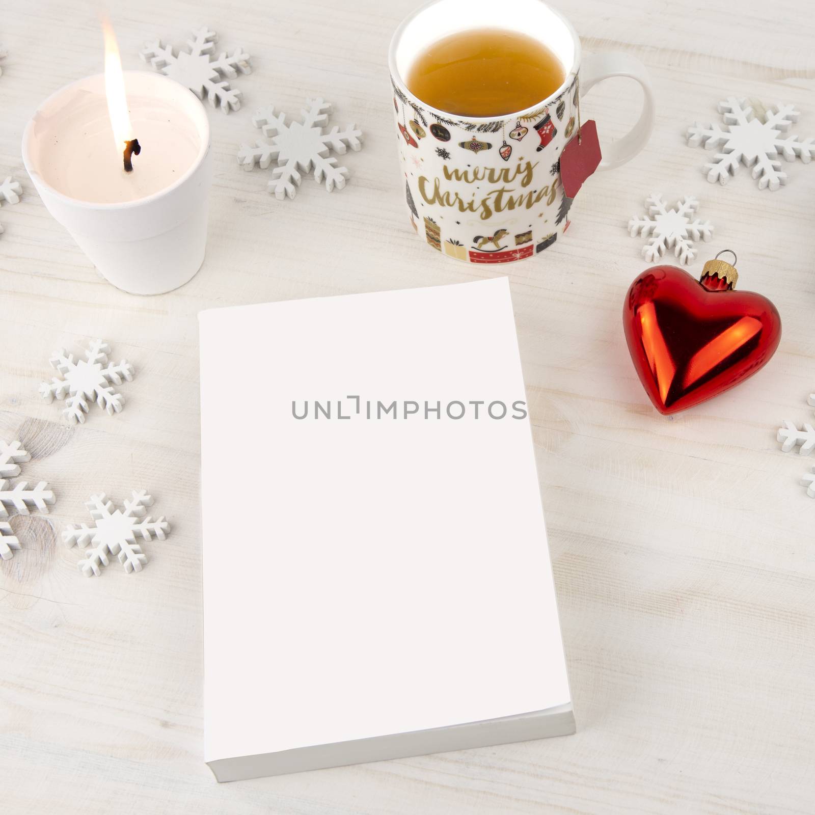 Christmas setting view from top for a book presentation: book with blank cover with a white lit candles, wooden snowflakes, Christmas cup of tea and red baubles with hearth shape on light wooden table by robbyfontanesi