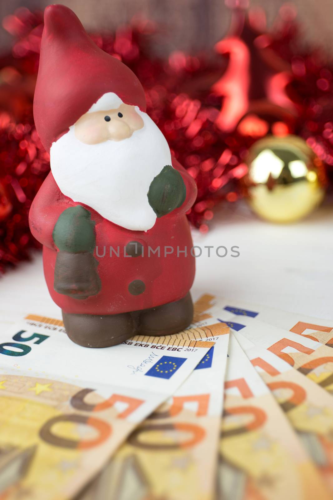 Christmas money business concept: a statuette of Santa Claus on some fifty euro banknotes with red and gold baubles and wreath decoration with bokeh effect on light wooden table