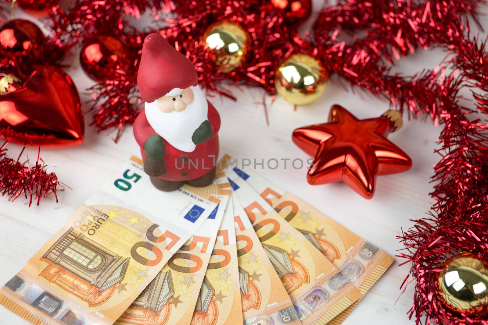 Christmas money business concept: a statuette of Santa Claus on some fifty euro banknotes with red and gold baubles and wreath decoration on light wooden table by robbyfontanesi