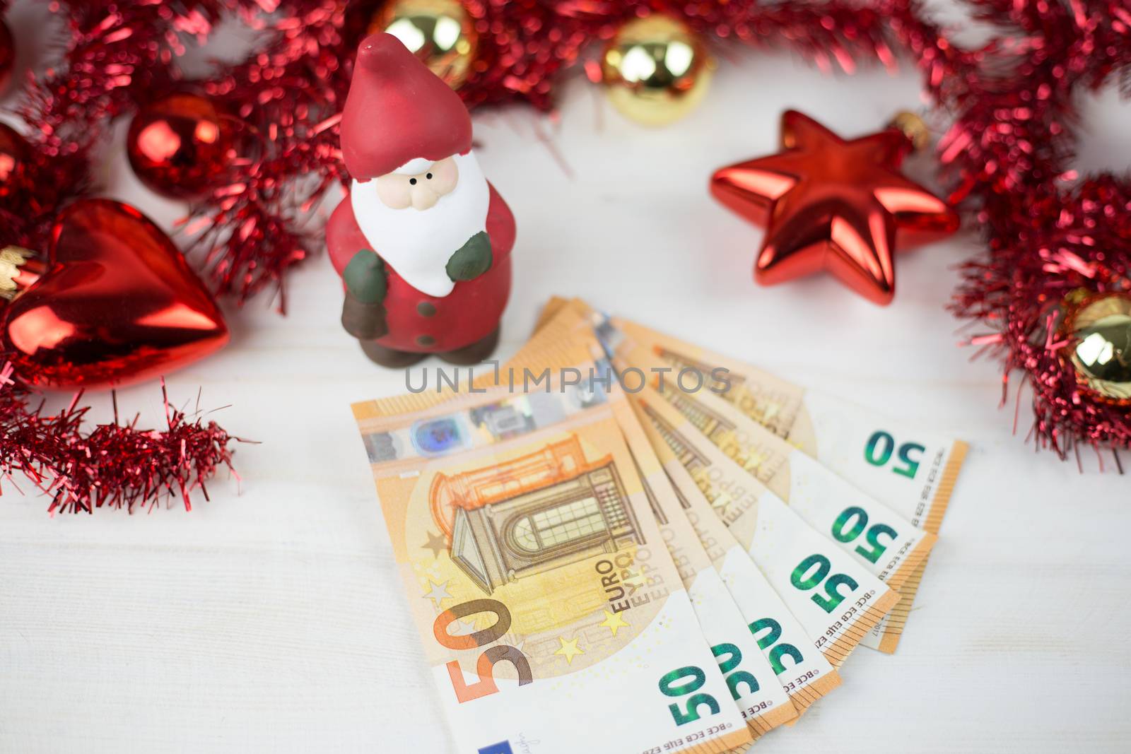 Christmas money business concept: a statuette of Santa Claus on some fifty euro banknotes with red and gold baubles and wreath decoration on light wooden table by robbyfontanesi