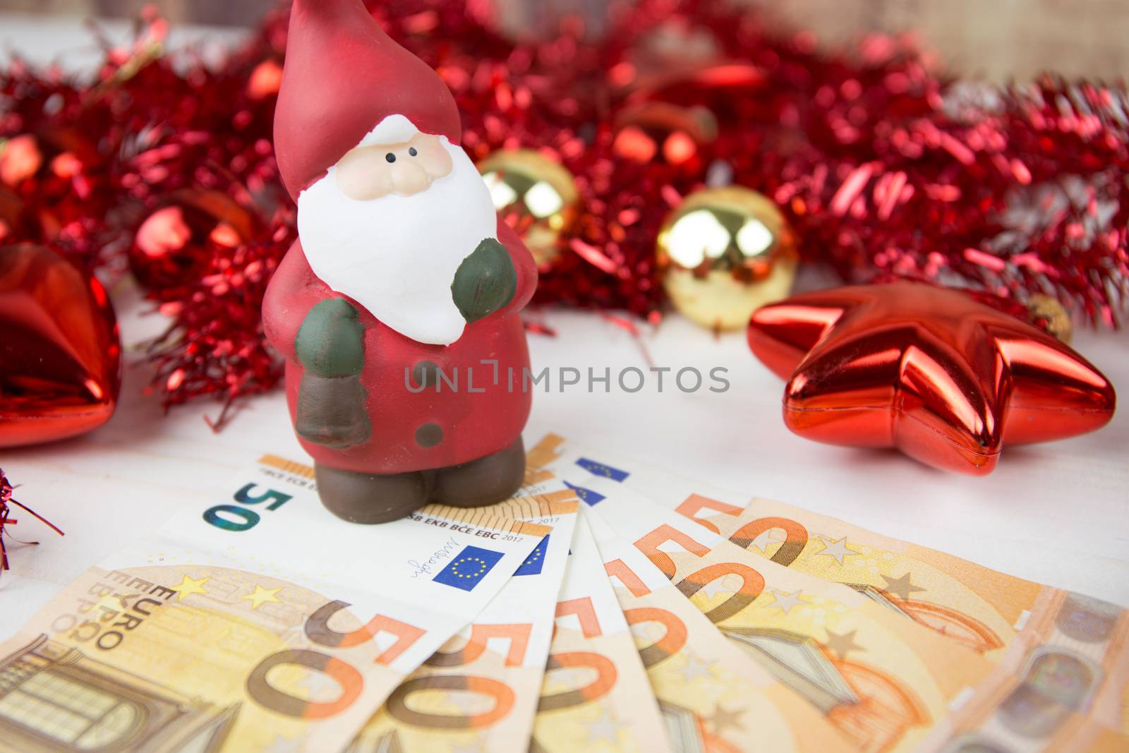 Christmas money business concept: a statuette of Santa Claus on some fifty euro banknotes with red and gold baubles and wreath decoration with bokeh effect on light wooden table by robbyfontanesi