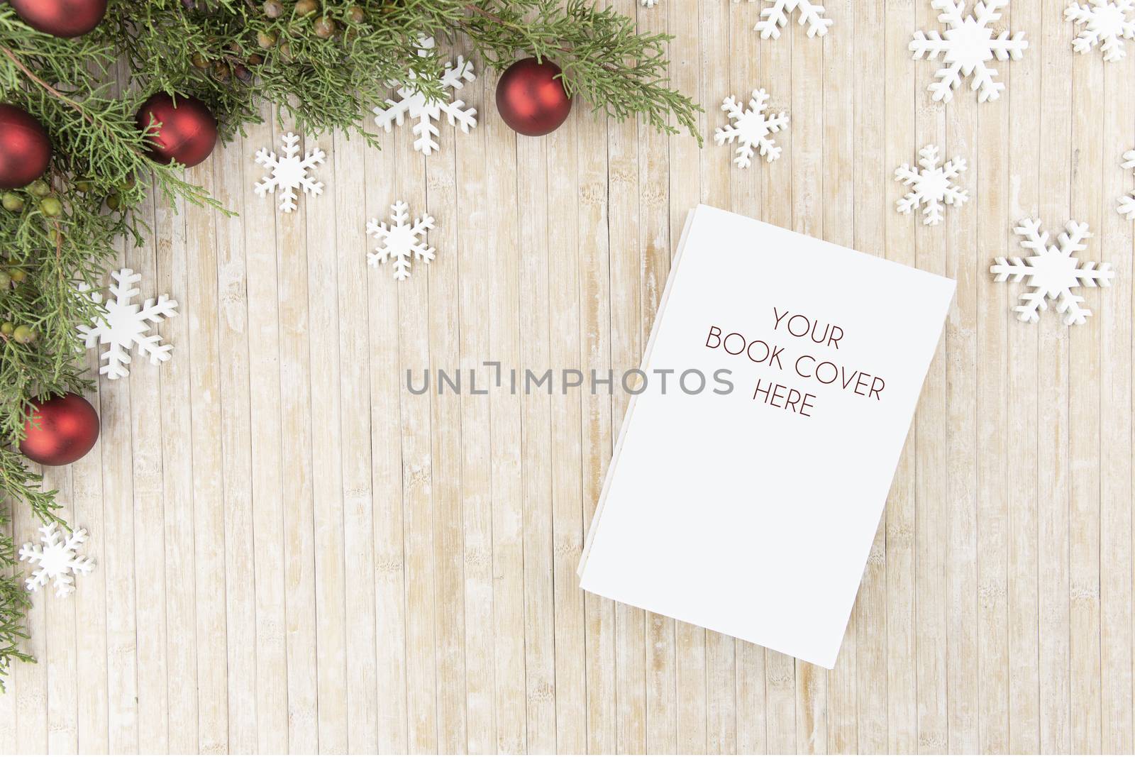 Christmas flat lay top view for a book presentation: book with blank cover, pine branches, white wooden snowflakes and red baubles on light wooden table by robbyfontanesi