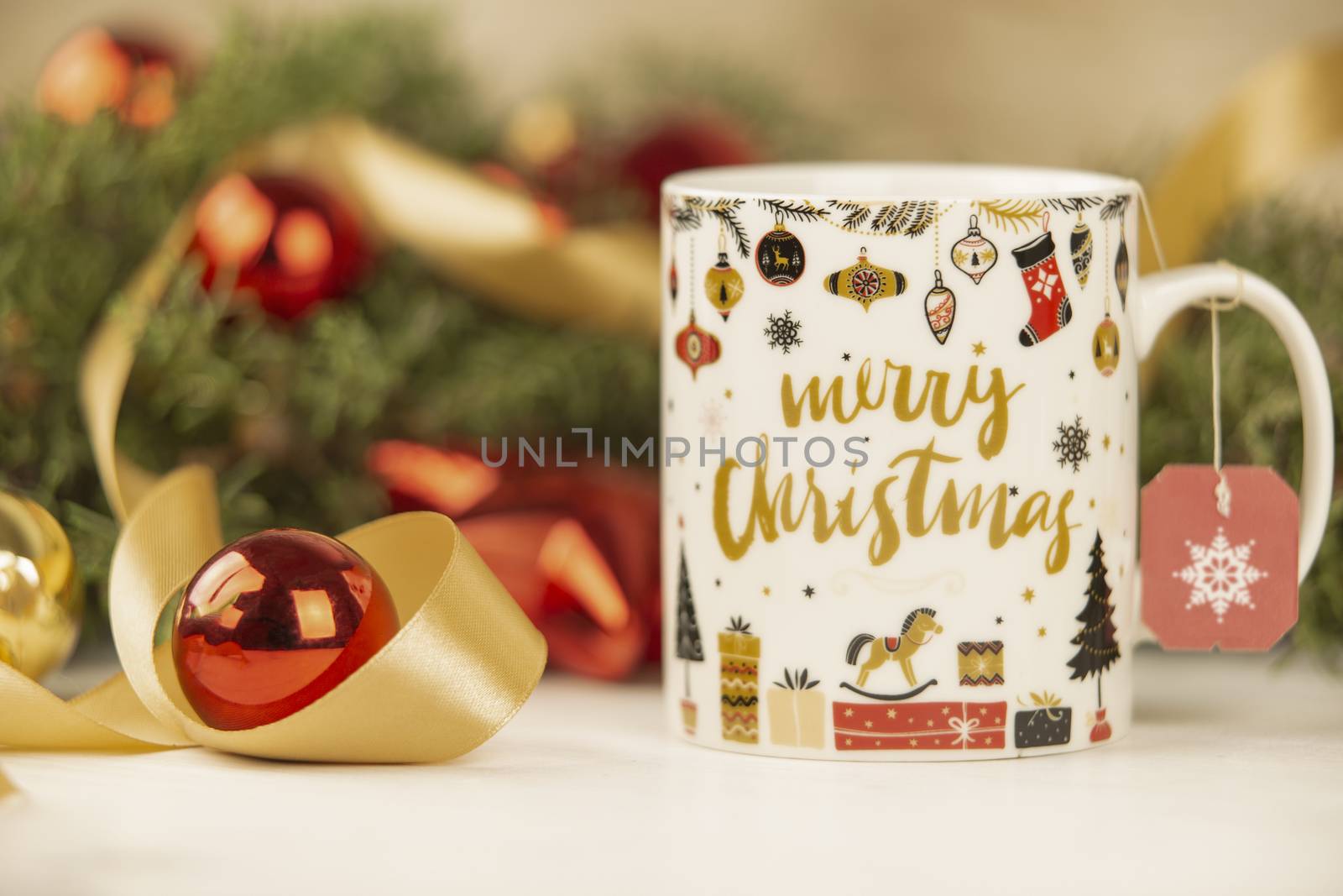 Close up of a Christmas mug with hanging empty tea label with around pine branches, red baubles and satin gold ribbon on wooden background with bokeh effect