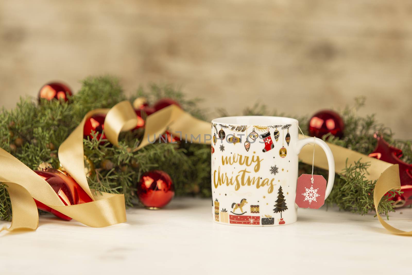 Close up of a Christmas mug with hanging empty tea label with around pine branches, red baubles and satin gold ribbon on wooden background with bokeh effect