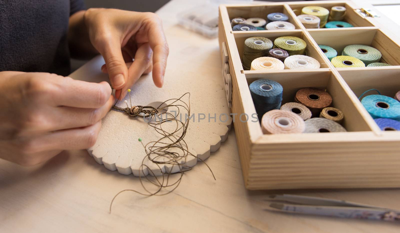 Lifestyle concept, work from home to reinvent your life: close-up of woman hands making macrame knotted jewelry with stone beads and tools on light wooden table by robbyfontanesi