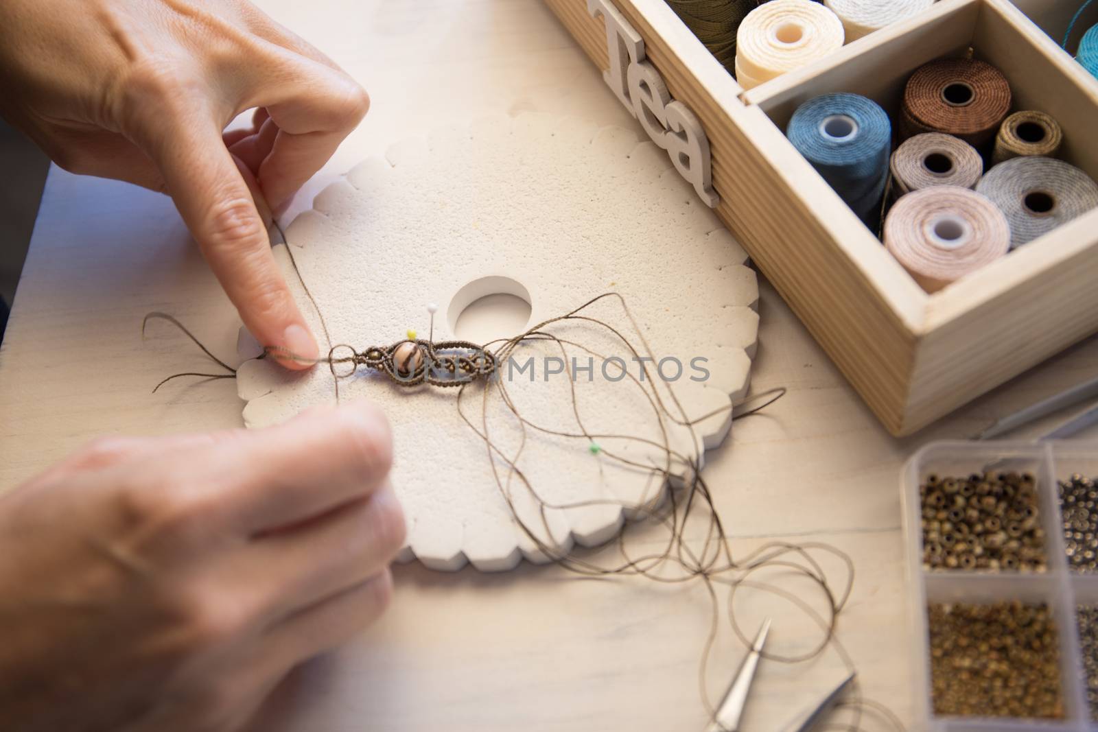 Lifestyle concept, work from home to reinvent your life: close-up of woman hands making macrame knotted jewelry with stone beads and tools on light wooden table