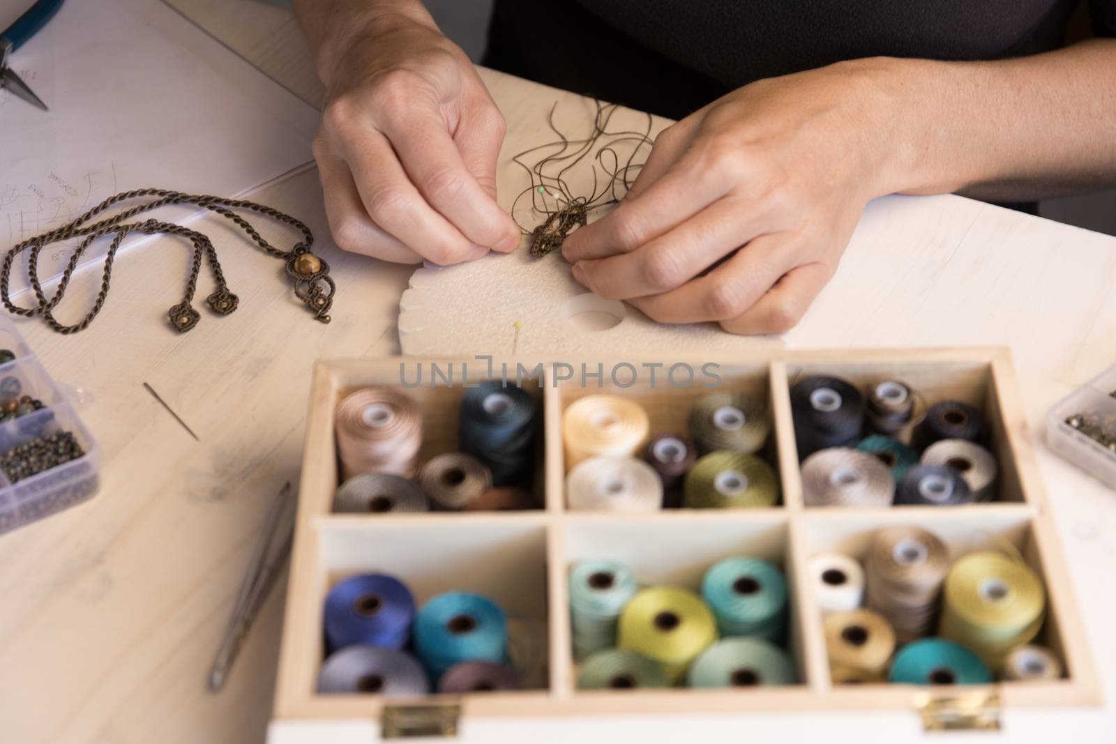 Lifestyle concept, work from home to reinvent your life: close-up of woman hands making macrame knotted jewelry with stone beads and tools on light wooden table