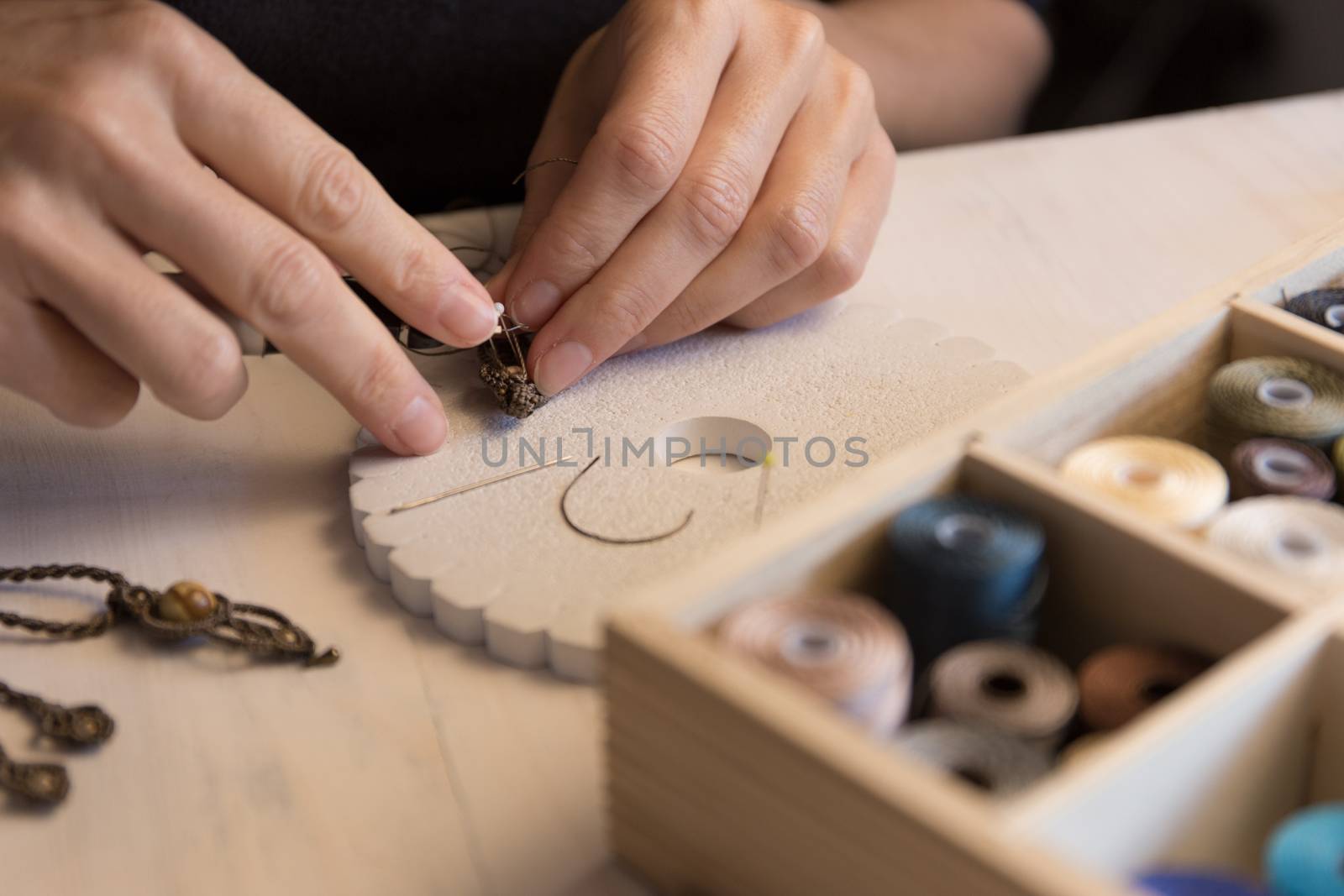 Lifestyle concept, work from home to reinvent your life: close-up of woman hands making macrame knotted jewelry with stone beads and tools on light wooden table