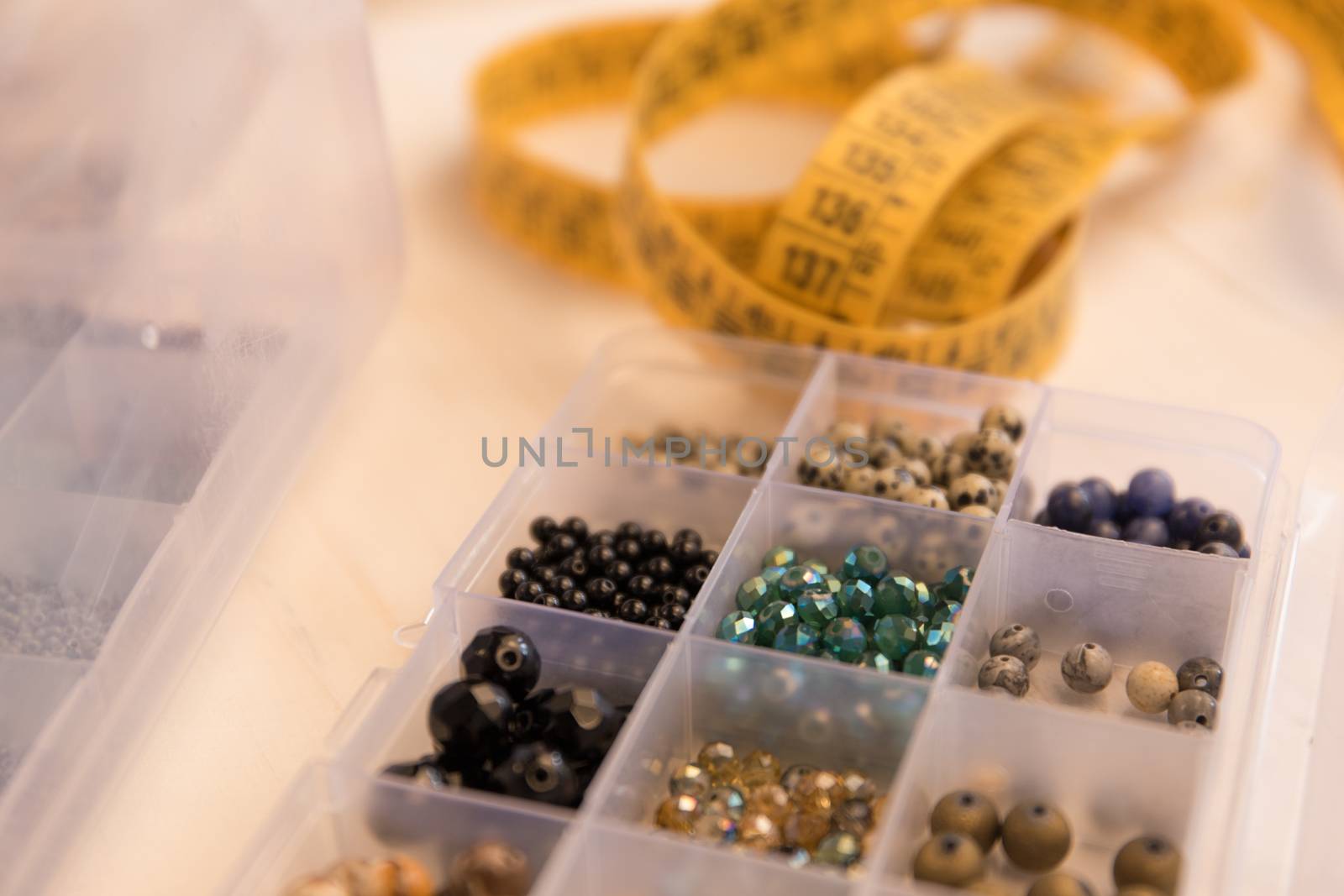 Work from home lifestyle concept: close-up detail of an organizer with various colored stone and crystal beads in foreground and a tailor meter in background bokeh effect on light wooden table
