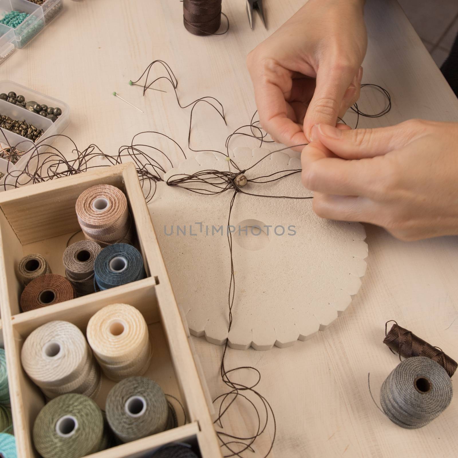 Lifestyle concept, work from home to reinvent your life: close-up of woman hands making macrame knotted jewelry with stone beads and tools on light wooden table