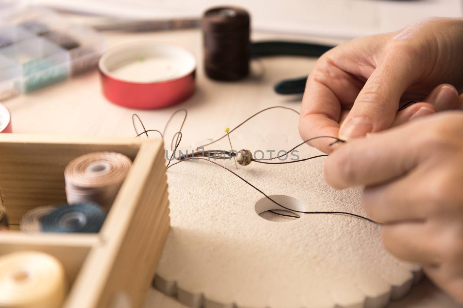 Lifestyle concept, work from home to reinvent your life: close-up of woman hands making macrame knotted jewelry with stone beads and tools on light wooden table