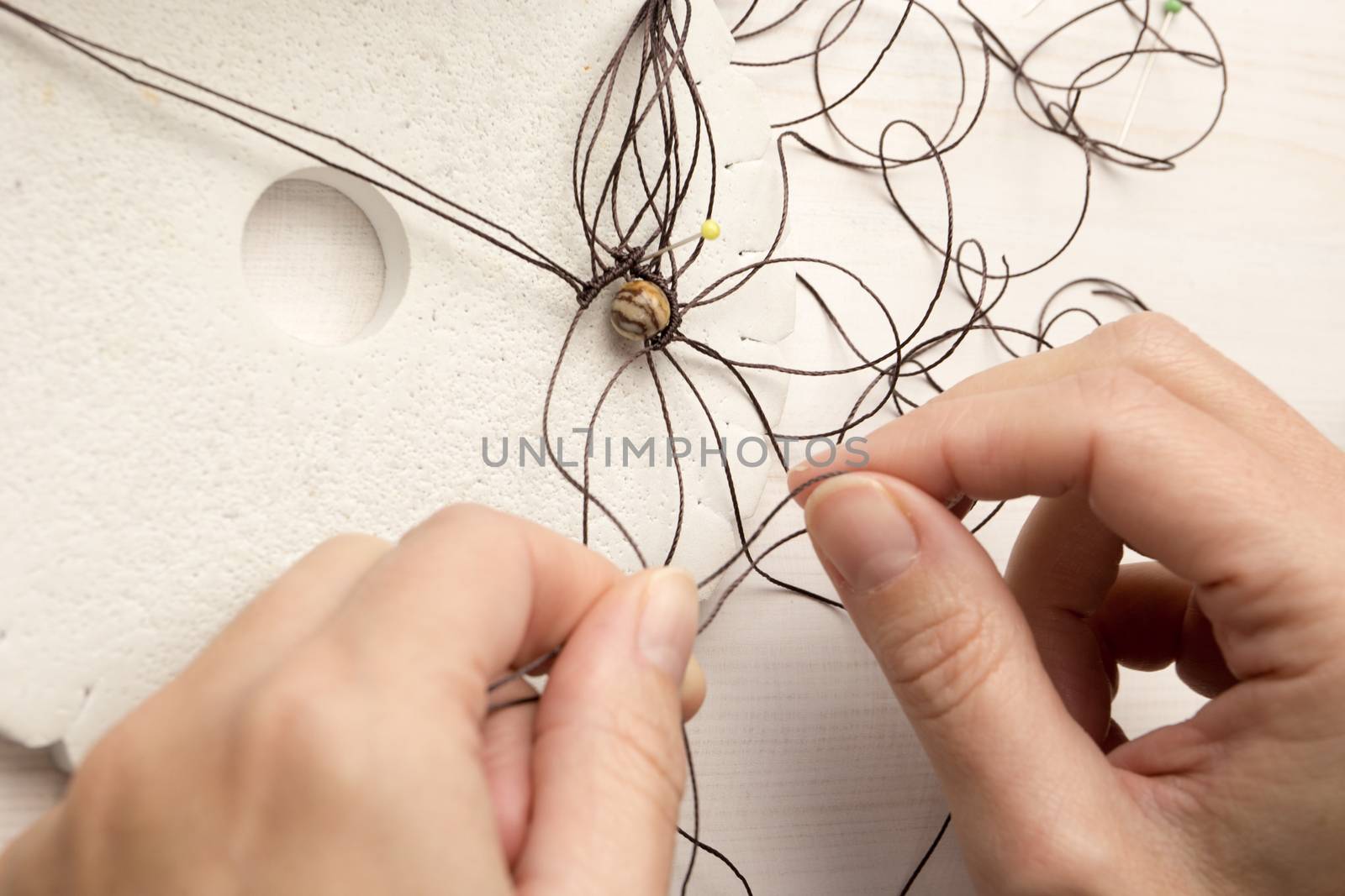 Lifestyle concept, reinvent your life and your job: close-up detail of woman hands making macrame knotted jewel with the fingers that tie the nylon thread around the diaspro natural stone