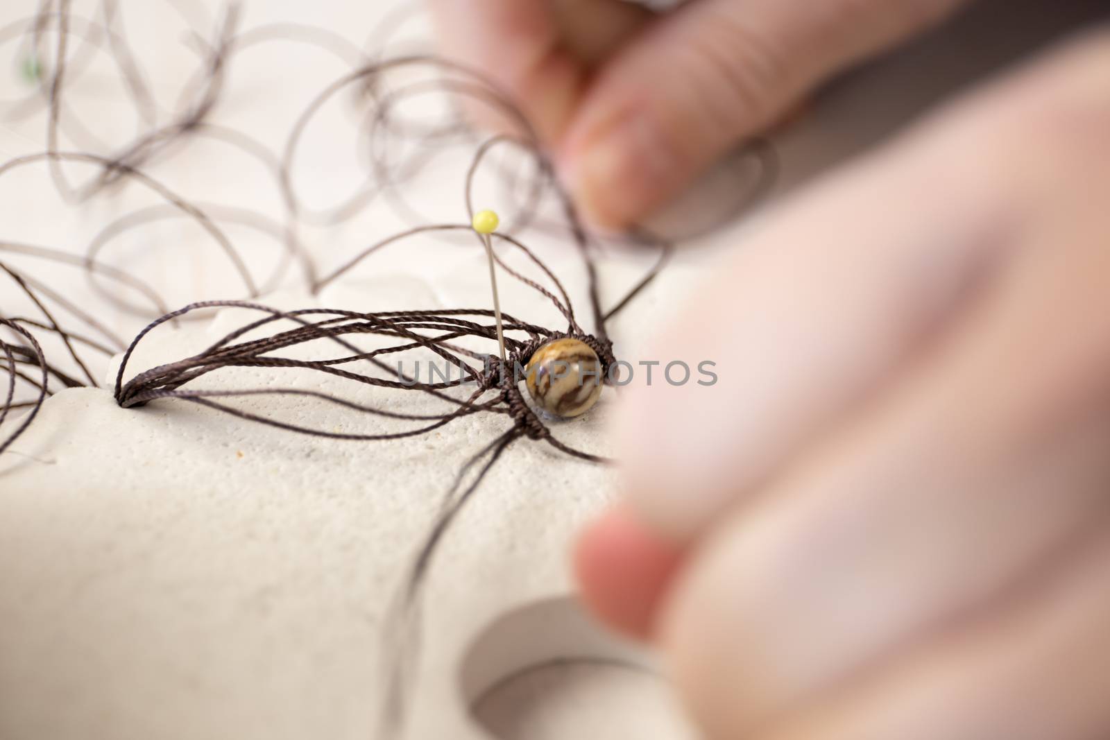 Lifestyle concept, reinvent your life and your job: close-up detail of woman hands making macrame knotted jewel with the fingers that tie the nylon thread around the diaspro natural stone by robbyfontanesi