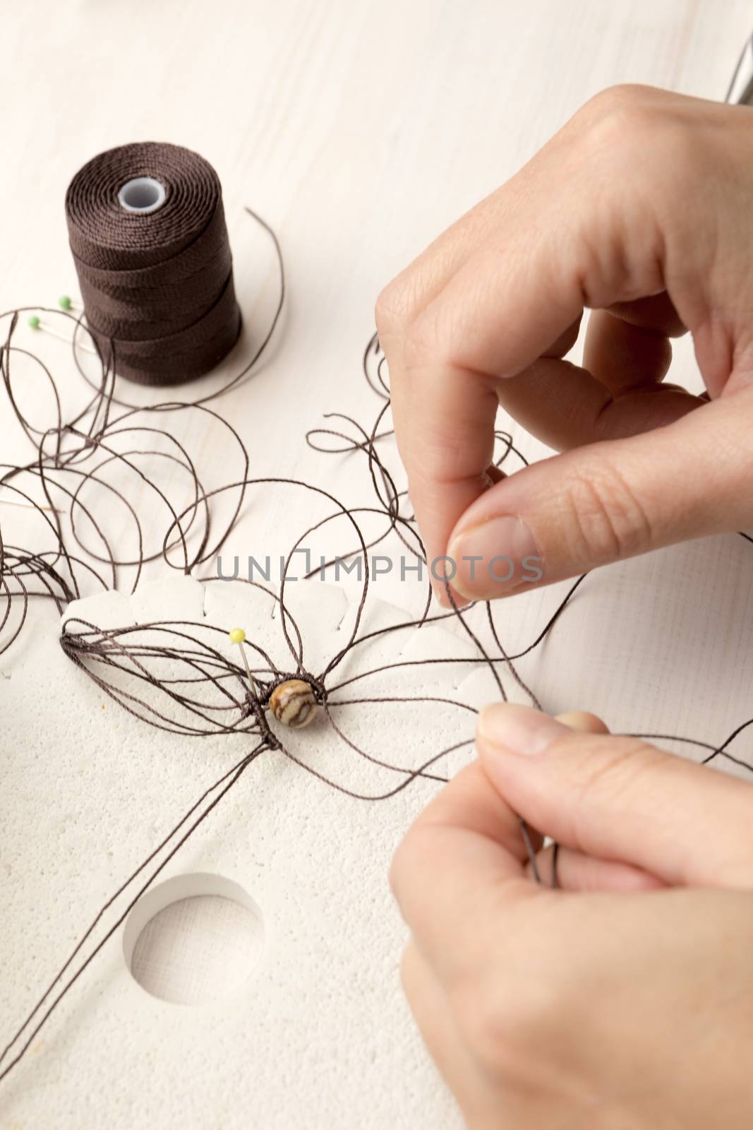 Lifestyle concept, reinvent your life and your job: close-up detail of woman hands making macrame knotted jewel with the fingers that tie the nylon thread around the diaspro natural stone