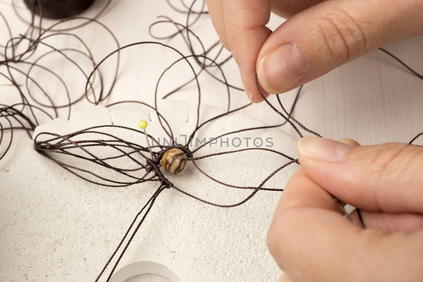 Lifestyle concept, reinvent your life and your job: close-up detail of woman hands making macrame knotted jewel with the fingers that tie the nylon thread around the diaspro natural stone