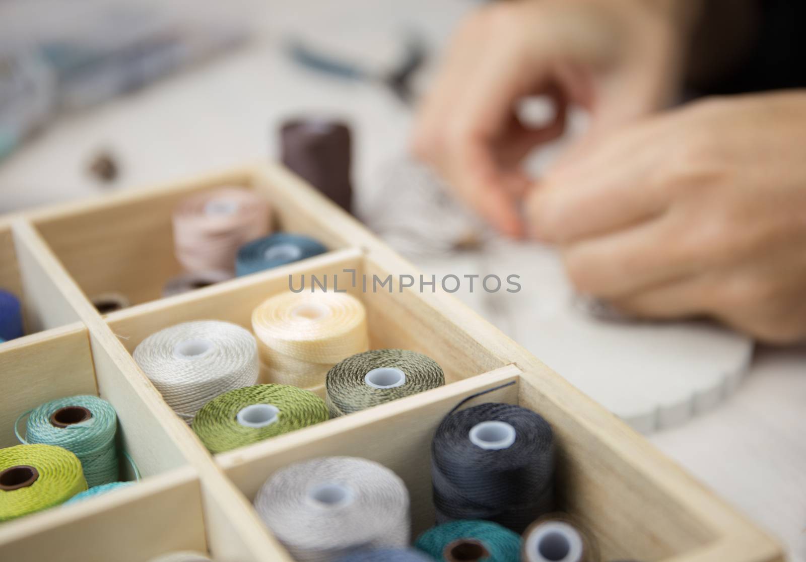 Lifestyle concept, work from home to reinvent your life: close-up detail of woman hands making macrame knotted jewel with some colored thread spools in foreground and bokeh effect by robbyfontanesi