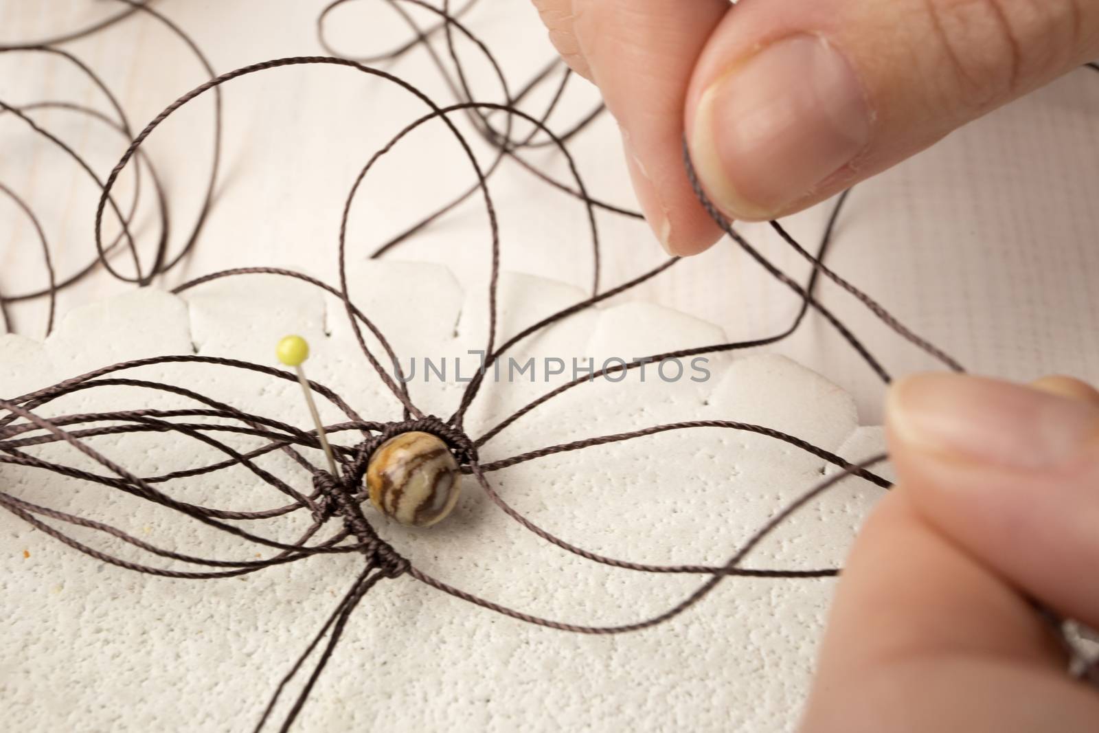 Lifestyle concept, reinvent your life and your job: close-up detail of woman hands making macrame knotted jewel with the fingers that tie the nylon thread around the diaspro natural stone