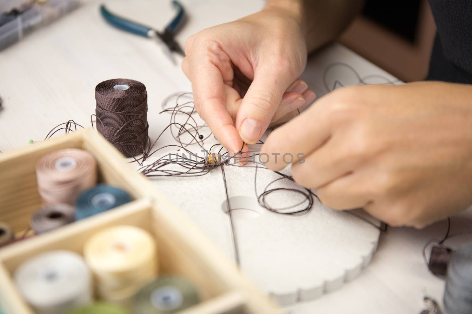 Lifestyle concept, work from home to reinvent your life: close-up detail of woman hands making macrame knotted jewel with some colored thread spools in foreground and bokeh effect