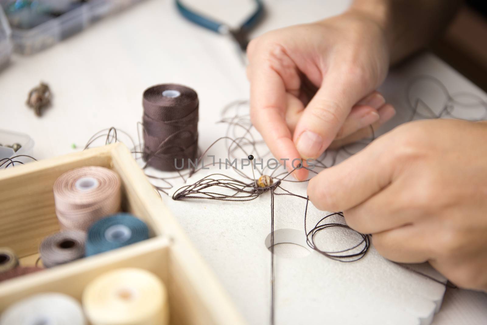 Lifestyle concept, work from home to reinvent your life: close-up detail of woman hands making macrame knotted jewel with some colored thread spools in foreground and bokeh effect by robbyfontanesi