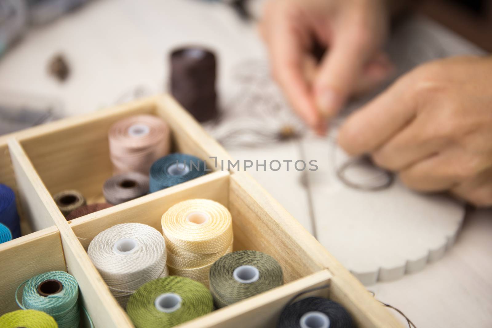 Lifestyle concept, work from home to reinvent your life: close-up detail of woman hands making macrame knotted jewel with some colored thread spools in foreground and bokeh effect by robbyfontanesi