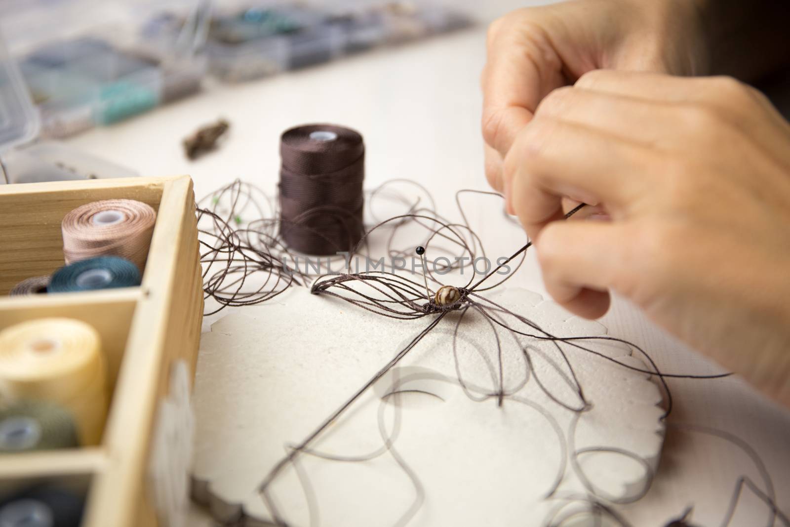 Lifestyle concept, work from home to reinvent your life: close-up detail of woman hands making macrame knotted jewel with some colored thread spools in foreground and bokeh effect by robbyfontanesi