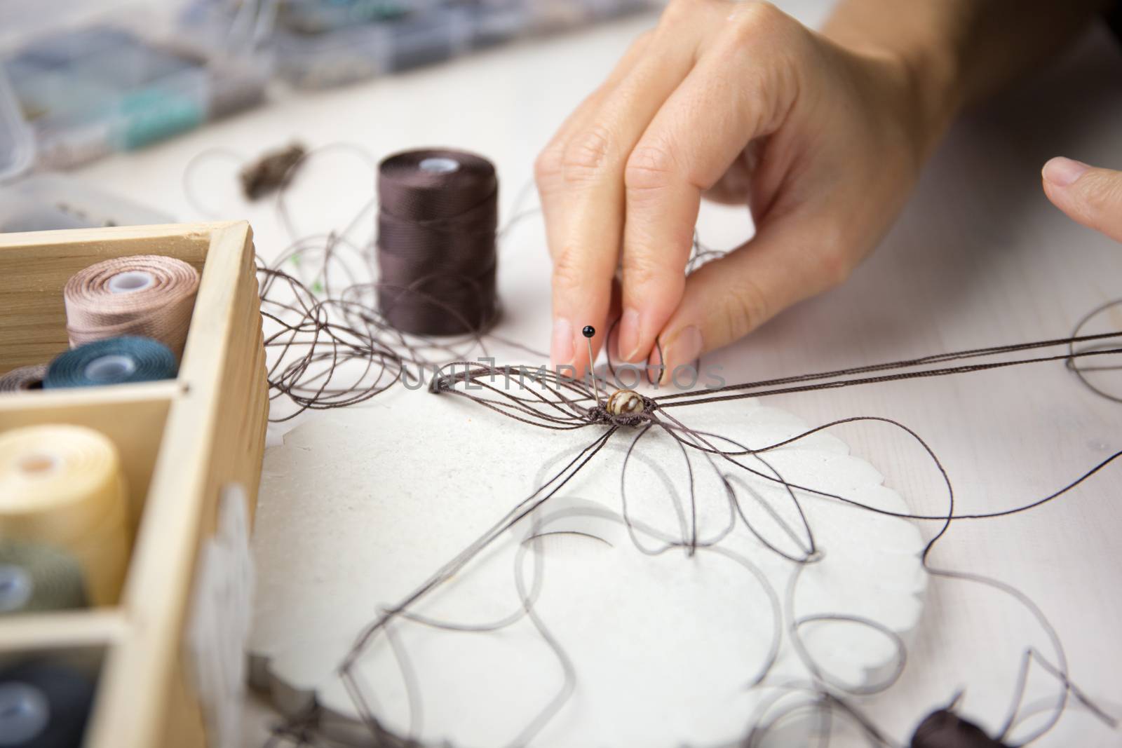 Lifestyle concept, work from home to reinvent your life: close-up detail of woman hands making macrame knotted jewel with some colored thread spools in foreground and bokeh effect by robbyfontanesi