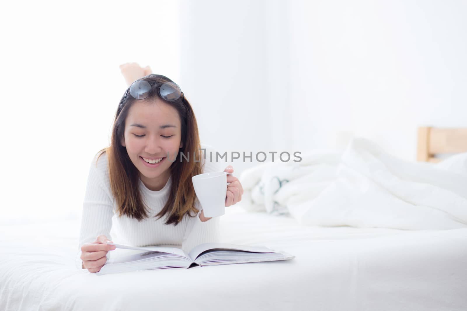 charming woman lying on bed reading a book and holding coffee cup looking into camera in bedroom.
