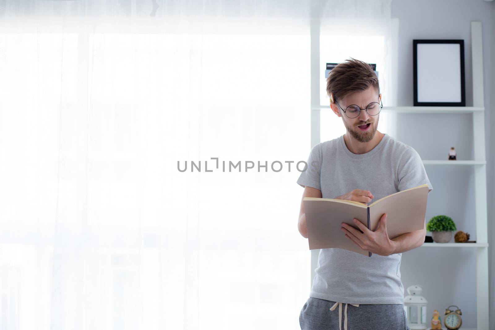 Handsome guy in eyeglasses is reading book preparing exam and thinking with standing at the living room - education concept.