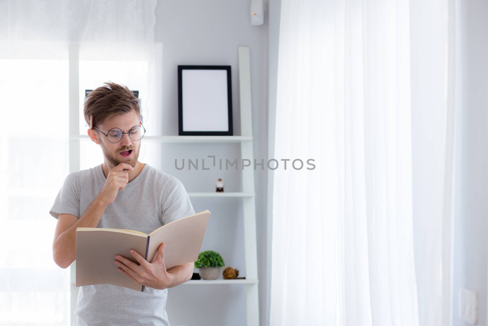 Handsome guy in eyeglasses is reading book preparing exam and thinking with standing at the living room - education concept.