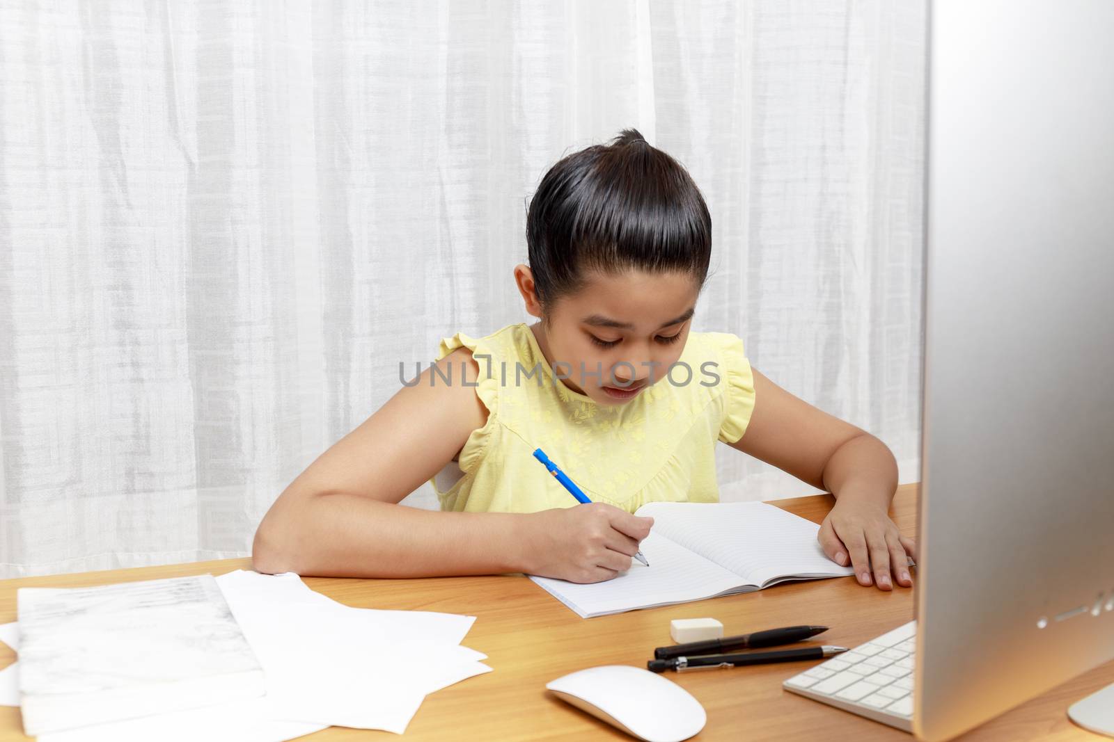 Asian little girl is studying online via the internet  sitting and writing in living room at home. Asia children writeing with pencil on notebook. online learning at home or learn frome home concept. 