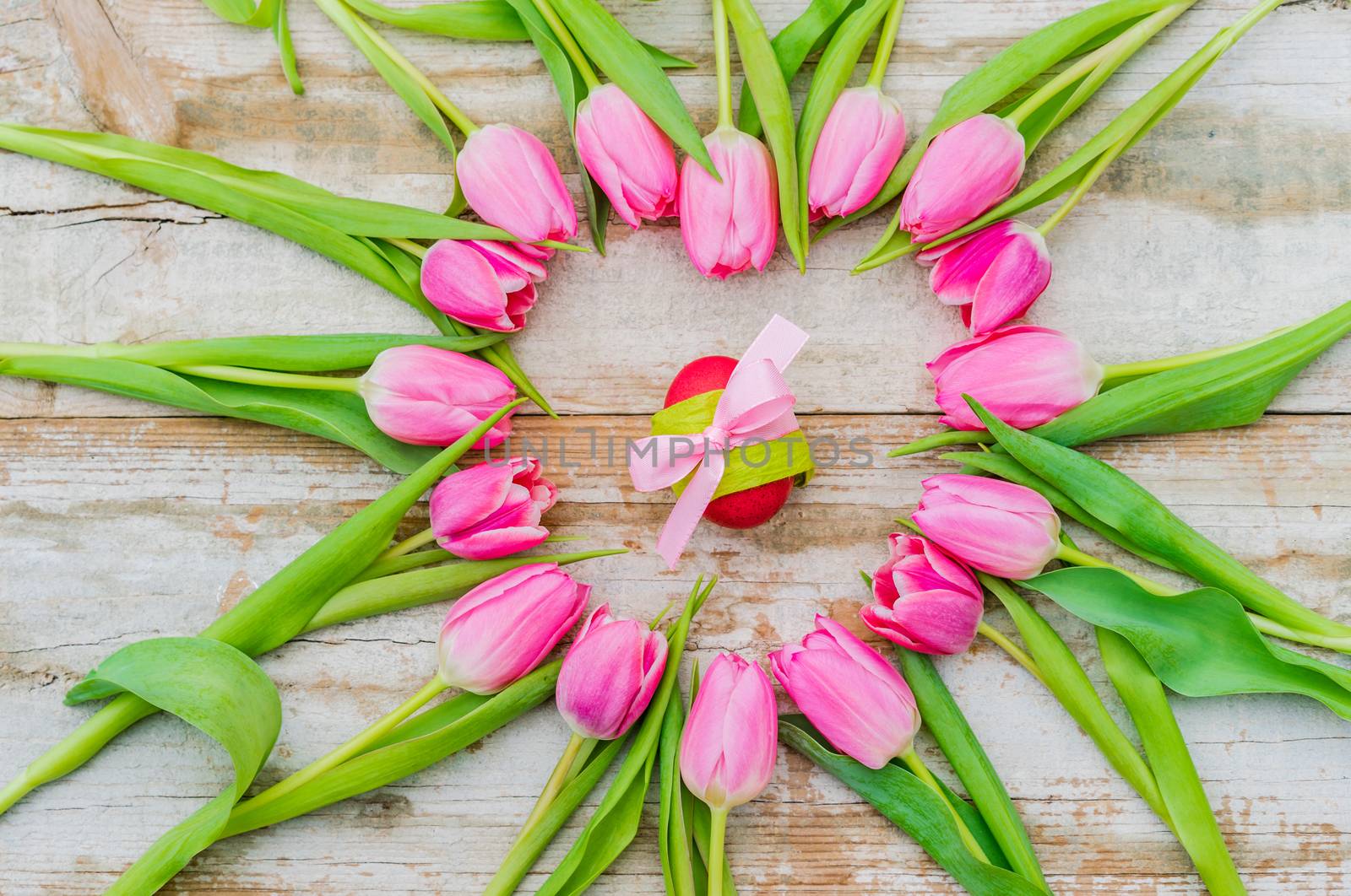 Easter decoration with egg ornated with pink ribbon bow, framed with pastel pink tulip flowers on rustic  wood