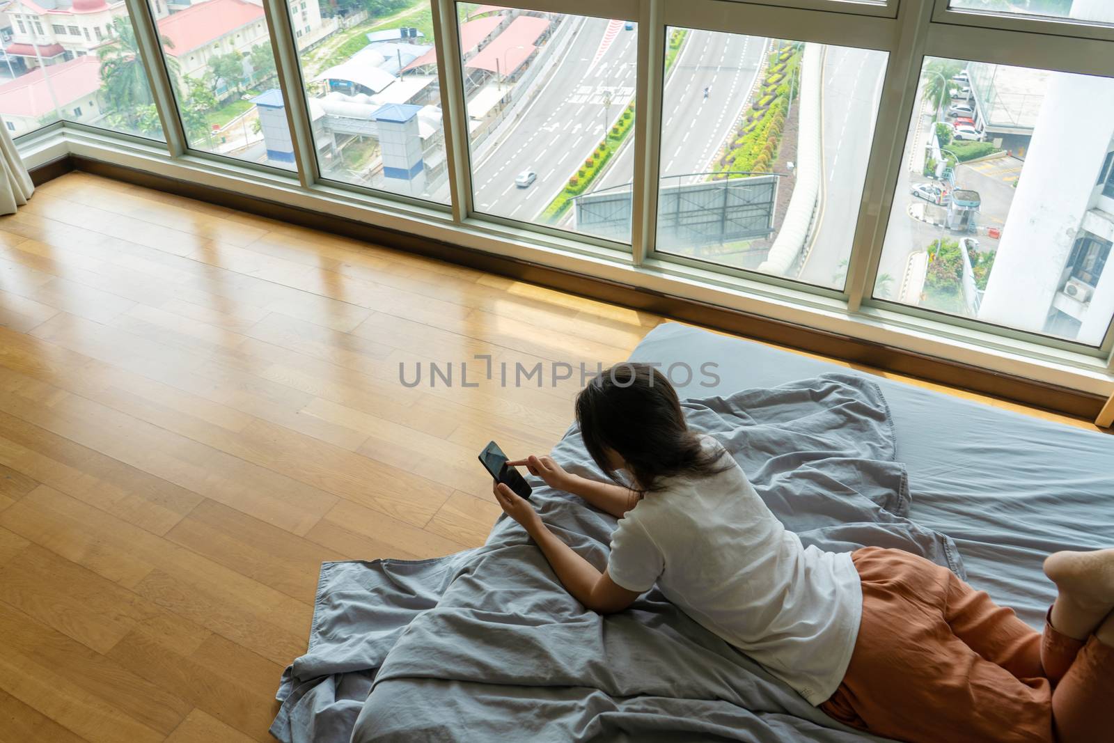 A beautiful young brunette girl is texting on the phone while lying on a bed by a panoramic window with a beautiful view from a high floor