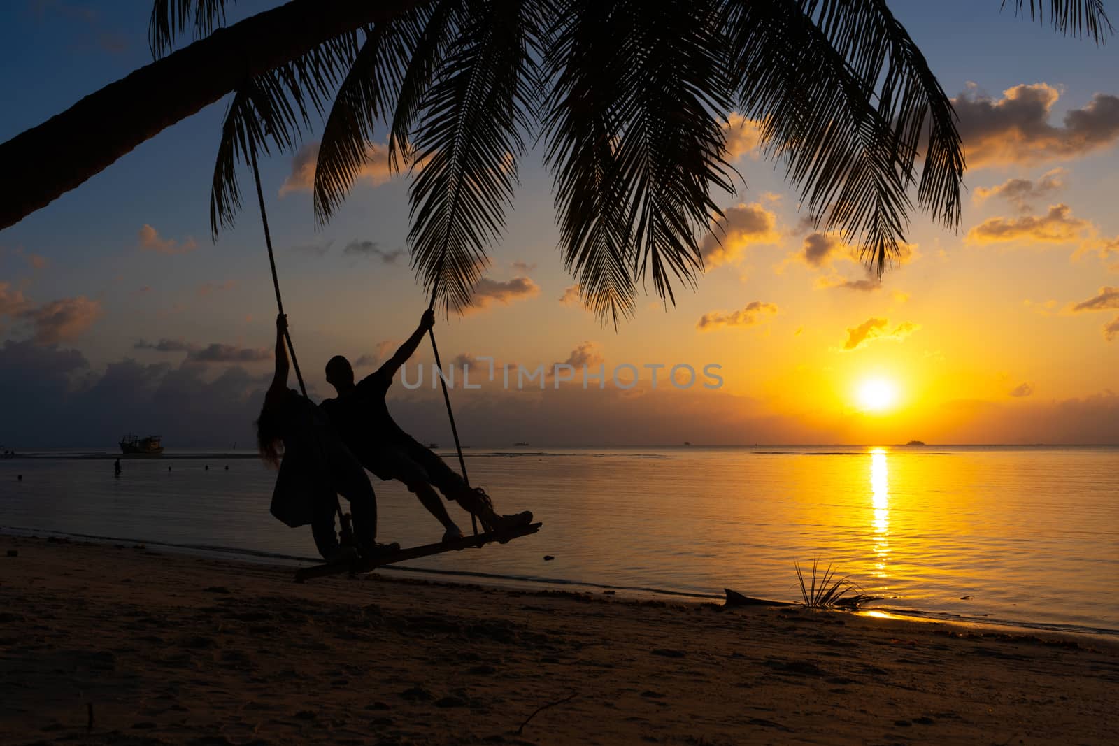 Silhouetted couple in love walks on the beach during sunset. Riding on a swing tied to a palm tree and watching the sun go down into the ocean by Try_my_best